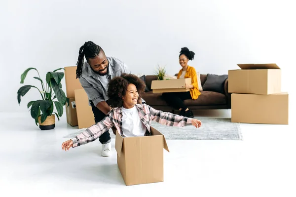 African-American family is having fun while moving to a new home, little daughter sits in a box, dad is pushing the box, laughing, having fun, mom sits on a couch, around cardboard boxes with things