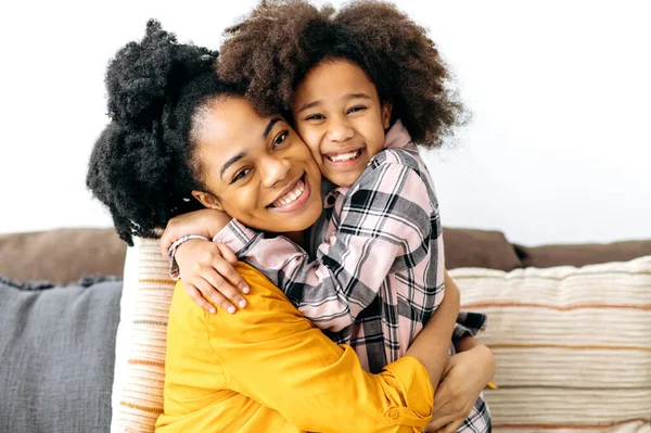 Happy mom and child. African American mom and her cute preschool daughter, hugging each other tightly while sitting on the sofa in living room at home, look at camera, smile. Love of parent and child
