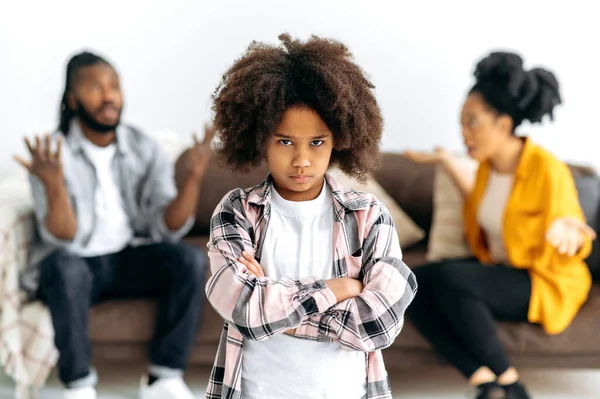 Difficulties in the family. An African American unhappy preschool girl looks offendedly at the camera, against the background, parents sort things out, shout at each other, swear, the child is scared