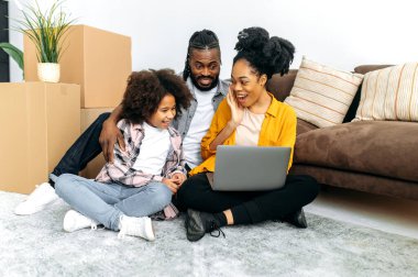 Cheerful happy African American married couple with preschool daughter, sit near a sofa in living room between cardboard boxes, using laptop,buy household goods in an online store, smile