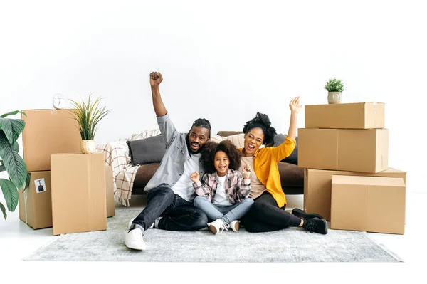 Cheerful happy African American married couple with preschool daughter, sit near a sofa in living room between cardboard boxes, rejoicing moving to new housing, look at camera, gesturing hands, smile