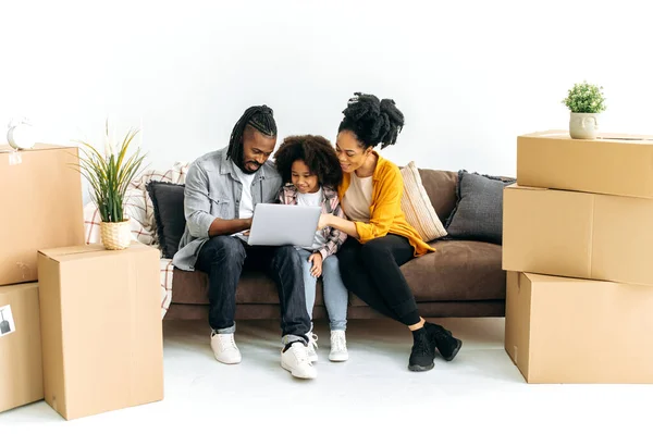 Happy African American family, dad mom and their preschool daughter, sit on a sofa in the living room between cardboard boxes, moved to new housing, using laptop,buy household goods in an online store