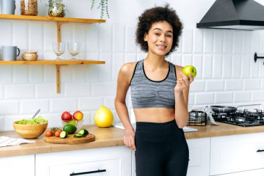 Slim beautiful mixed race curly haired girl in sportswear stands at home in the kitchen, holds an apple in her hand, prepares a healthy lunch, looks at the camera, smiles. Healthy food concept