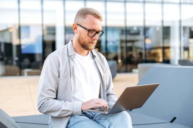 Working in a laptop. Concentrated caucasian man with a glasses, in a casual clothes, IT specialist, developer, programmer, working in a laptop outdoors, developing a new program, writing code