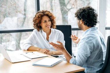 Two positive confident motivated employees of the company of different nationalities are working together on a new project in a modern office, together discussing ideas and goals, communicating