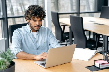 Focused smart confident guy of Indian nationality with curly hair, a programmer, IT specialist, sits at a table in the office, works in a laptop, works on a new program, at software support
