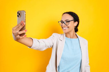 Neşeli, kendine güvenen, gözlüklü, şık giyinmiş, cep telefonunu elinde tutan, telefon kamerasına bakan, selfie çeken, gülümseyen, izole edilmiş sarı arka planda duran güzel Brezilyalı ya da Latin bir kadın.