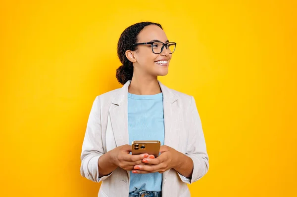 stock image Lovely confident mixed race woman with glasses, stylishly dressed, using mobile phone, chatting in social media, texting message, looks to the side, smiling, stand on isolated yellow background