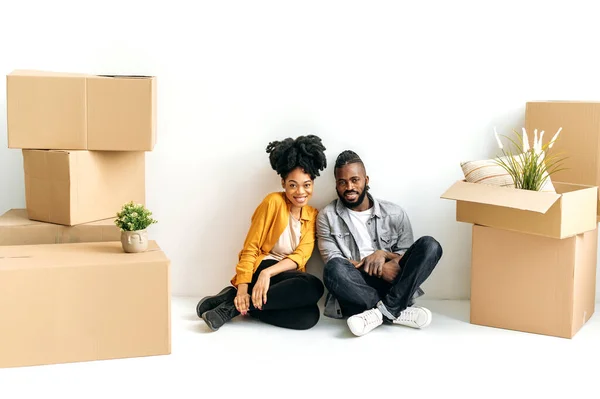 快乐的非洲裔美国夫妇 身穿休闲装 坐在地板上自己的第一座房子里 身边放着装有东西的纸盒 他们看着相机 微笑着 投资于房地产 — 图库照片