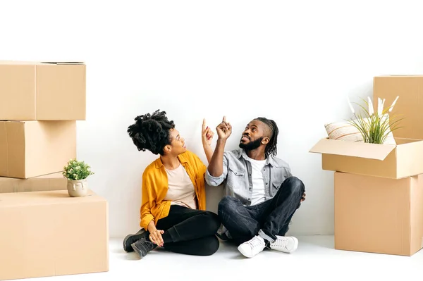 快乐的非洲裔美国人夫妇坐在地板上的纸板箱与家居用品 计划修复他们的第一个家 规划新公寓的未来室内设计 — 图库照片