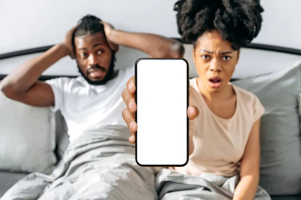 Shocked puzzled african american couple, sit in a cozy bed in the bedroom, woman shows smartphone with blank mock-up screen, space for advertising, presentation, look at camera at a loss