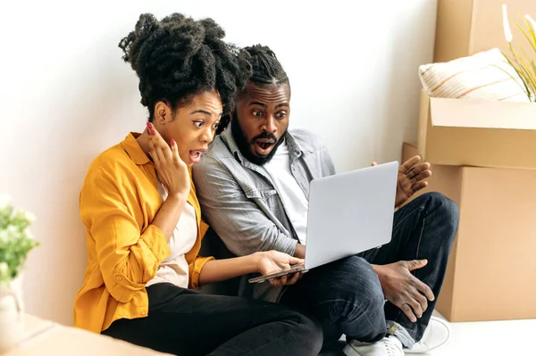 Shocked stunned african american married couple, dressed in casual wear, sit on the floor between cardboxes with things around, they use laptop for online shopping, look in amazement at the screen