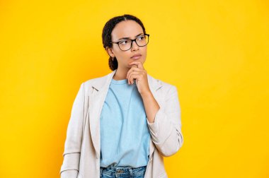 Endişeli, melez, genç, esmer iş kadını, elini çenesine yakın tutuyor, düşünceli bir şekilde yan yana bakıyor, hayal kuruyor, planlıyor, düşünüyor, izole edilmiş sarı arka planda duruyor.