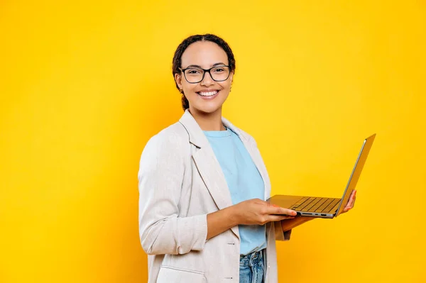 stock image Positive confident successful brazilian or hispanic business woman, company employee, consultant, with glasses, hold in hands open laptop, stand on isolated yellow background, looks at camera, smiling