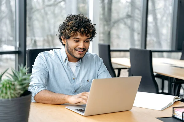 Positive satisfied Indian man, company employee, executive, sits at a work desk in the office, working in a laptop on a new project, financial business plan, strategy, looks at laptop screen, smiles