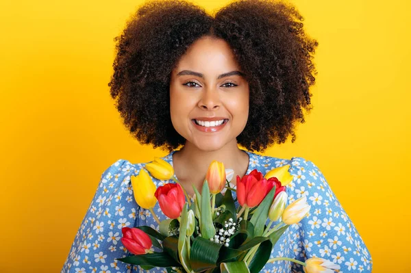 Happy lovely curly haired african american young woman in a blue summer dress, holding bouquet of colorful tulips on womens day or birthday, stand on isolated yellow background, looks at camera,smile