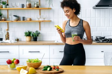 Sağlıklı gıda konsepti. Pozitif ince Brezilyalı ya da Latin bir kız, mutfakta durur, salata hazırlar, akıllı telefon kullanır, yemek tarifi arar, elinde bir bardak taze yeşil smoothie taşır.
