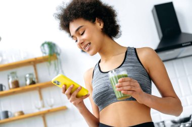 Healthy eating, veganism, raw food. Pretty, happy, healthy mixed race woman, stand in a kitchen, using smart phone, holding a glass of healthy fortified organic smoothie, taking care of health, smiles