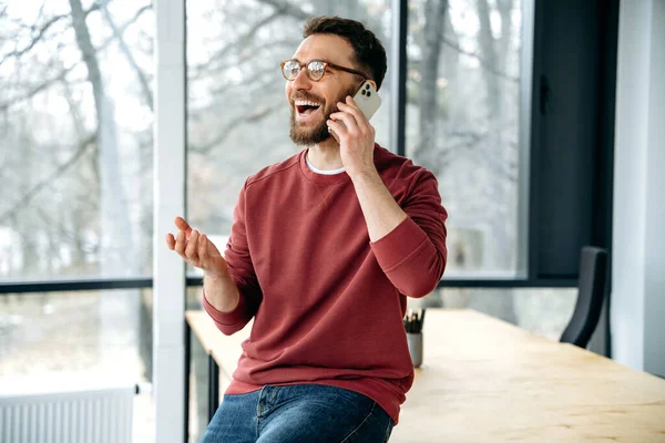 Smart phone conversation. Successful caucasian man, office manager, having a work call while sitting on a table in the office, consulting clients on phone, discuss project, looking away, smiling