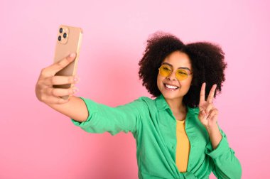 Güzel esmer Brezilyalı kadın, yeşil günlük gömlek giyiyor, akıllı telefon kullanıyor, selfie çekiyor, parmaklarıyla barış hareketi gösteriyor, kamera ekranına bakıyor, gülümsüyor, izole edilmiş pastel pembe arka plan