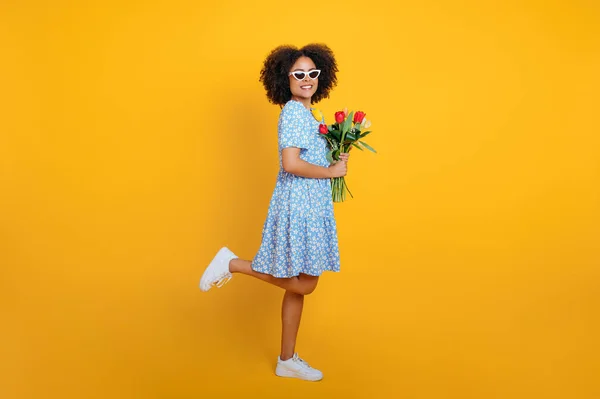 Full length photo of happy lovely curly brazilian woman in blue summer dress, holding bouquet of colorful tulips on womens day or birthday, stand on isolated orange background, looks at camera, smile