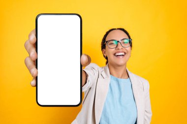 Heyecanlı mutlu Brezilyalı ya da gözlüklü İspanyol kadın, anonsu için beyaz ekranlı akıllı telefonunu gösteriyor, kameraya bakıyor, gülümsüyor, izole edilmiş sarı arka planda duruyor.