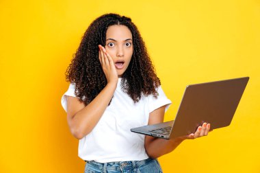 Şaşırmış Brezilyalı ya da İspanyol kadın, kıvırcık saçlı, beyaz, basit tişörtlü, elinde açık bir laptop tutan, şaşırmış bir şekilde kameraya bakan, izole edilmiş sarı arka planda duran