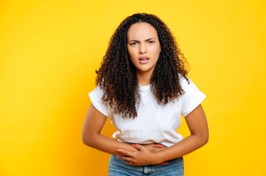 Brezilya 'lı ya da İspanyol asıllı genç bir kadın, günlük giysiler giymiş, karın ağrısı, spazm, regl sırasında ağrı çekiyor, ellerini karnının üstünde tutuyor, izole edilmiş sarı arka planda duruyor.
