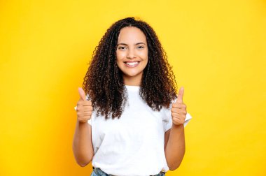 Güzel, pozitif Brezilyalı ya da İspanyol kıvırcık saçlı, sıradan beyaz tişörtlü, kameraya gülümseyen, iki eliyle hareket eden, izole edilmiş sarı arka planda duran