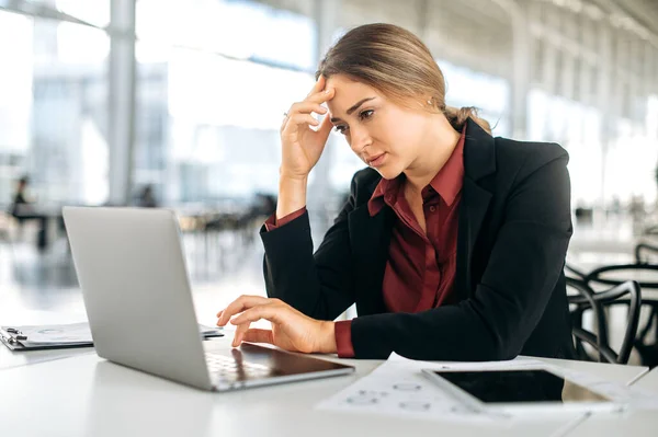 Stressed exhausted frustrated worried caucasian woman, company employee, product manager, sits at her work desk, sadly looks at laptop screen, feels tired from work, has problems with the project