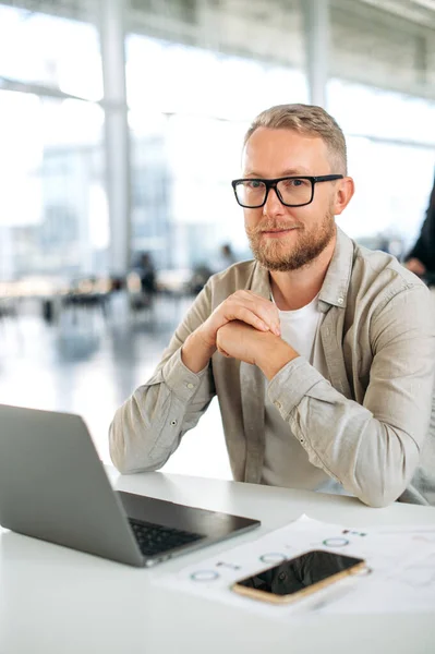 Vertical photo of a positive attractive clever caucasian business man, with glasses, programmer, company seo, software administrator, sits at a desk with a laptop in the office, looks at camera, smile