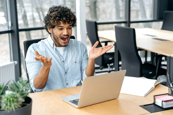 Happy excited indian or arabian businessman, company employee, manager, sitting at workplace in the office, looks in amazement at a laptop screen, received unexpected news, rejoices in success, smile