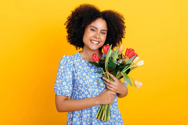 stock image Charming positive african american woman with curly hair, wearing summer blue dress, holding bouquet of colorful tulips, received gift, looks at camera, smiling, stands over isolated orange background