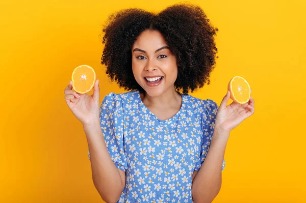 Beautiful excited african american curly haired woman in a summer blue dress, holding two halves of citrus juicy fruits in hands, posing and smiling at camera, stand on isolated orange background