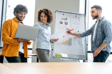 Toplantı odasında grup beyin fırtınası. Bir grup pozitif çok ırklı meslektaşlar, modern ofiste beyaz tahtaya yakın durun, fikirler hakkında konuşun, riskleri analiz edin ve kazancı analiz edin, gülümseyin.