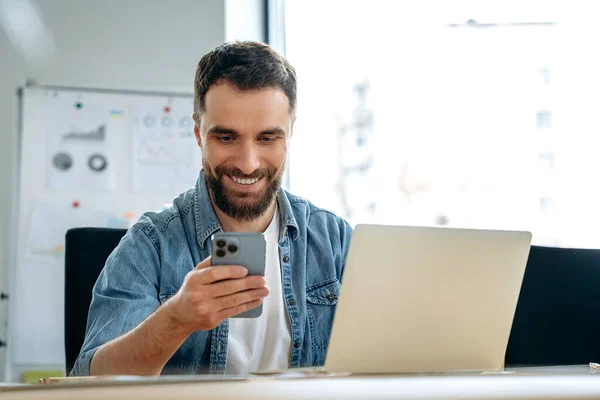 Satisfied caucasian man with beard, freelancer, programmer, seo, sits at a table in the office, uses his smartphone, messaging on social networks, conducts work correspondence, watches news, smiles