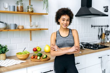 Güzel, zayıf, Afro-Amerikan spor kıyafetli genç bir kadının fotoğrafı. Mutfakta kollarını kavuşturmuş, taze salata hazırlarken kameraya bakıyor, dostça gülümsüyor.