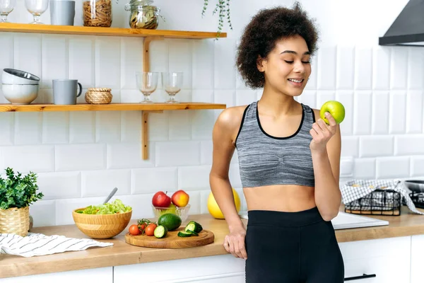 stock image Healthy food concept. Lovely healthy african american curly haired young woman in sportswear stands at home in the kitchen, holds an apple in her hand and looks at it, prepares a healthy lunch, smiles