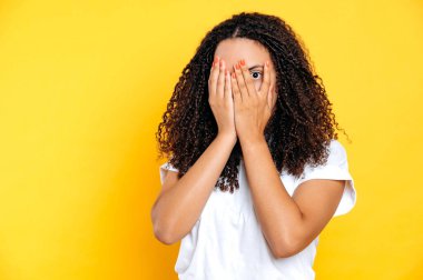 Korkmuş İspanyol ya da Brezilyalı kıvırcık saçlı beyaz tişörtlü kadın, parmaklarının arasından bakıyor, yüzünü ellerle örtüyor, korku yaşıyor, şok geçiriyor, sarı arka planda duruyor.