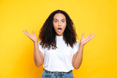 Şaşırmış Brezilyalı ya da beyaz tişörtlü İspanyol kıvırcık kadın kameraya şaşkınlıkla bakıyor ağzı açık, şaşırmış, elleriyle işaret ediyor, izole edilmiş sarı arka planda duruyor.