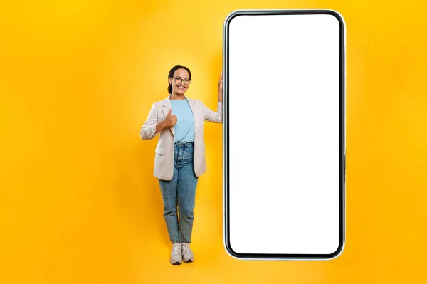 Full length photo of brazilian woman, stand near an enlarged mobile phone with blank white mock-up screen for presentation or advertising, shows thumb-up at camera, smile, yellow isolated background
