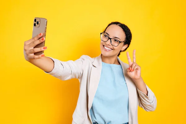 stock image Joyful lovely pretty brazilian woman with glasses, stylishly dressed, holds smart phone in hand, looks at phone camera, takes a selfie photo, smile and show peace gesture, isolated yellow background