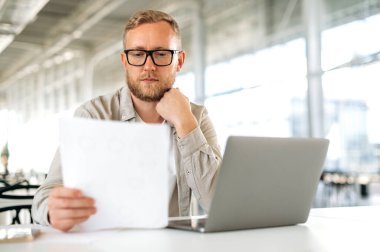 Odaklanmış beyaz adam, şirket çalışanı, ürün yöneticisi, çalışma masasında oturur, belgelere dikkatle bakar, bilgi analiz eder, sonuç çıkarır, planlama stratejisi ve promosyon yapar.