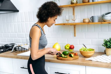 Sportif ince, sağlıklı Afro-Amerikan kıvırcık saçlı, spor kıyafetli kadın mutfakta taze salata için sebze kesiyor, gülümsüyor. Vejetaryenlik, çiğ gıda, sağlıklı organik gıda kavramı.