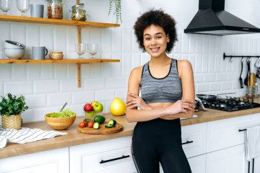 Afro-Amerikan kıvırcık saçlı güzel bir kadının fotoğrafı, spor kıyafetleri giymiş, kollarını kavuşturup mutfakta duruyor, kameraya bakıyor, gülümsüyor, salata için taze sebzeler arka planda yatıyor.