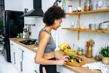Sağlıklı yemek hazırlığı. Mutfakta salatalık keserken mutlu bir şekilde gülümseyen spor kıyafetli, sağlıklı, zayıf, Afro-Amerikan kıvırcık kadın.