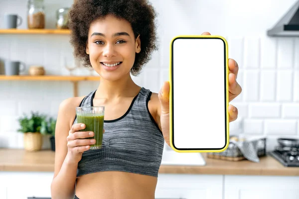 Positive smiling pretty african american woman holding a glass of a fortified organic green smoothie and showing smart phone with empty white mock up screen for the recipe, presentation, advertising