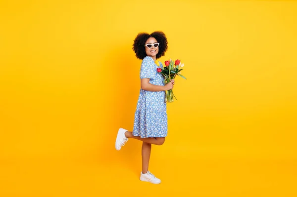 Full length photo of gorgeous curly brazilian woman in blue summer dress, holding bouquet of colorful tulips on womens day or birthday, stand on isolated yellow background, looks at camera, smiles