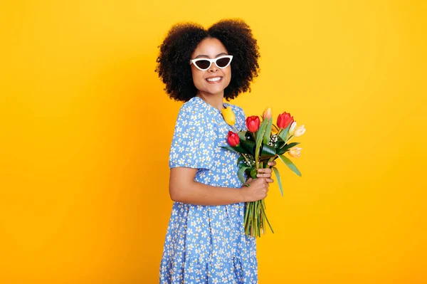 Fashionable brazilian or hispanic woman with glasses, in a blue summer dress, holding a bouquet of fresh multicolored tulips for a birthday or womens day, smile at camera, isolated orange background