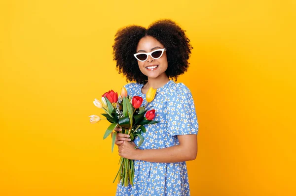 Portrait of fashionable african american woman with glasses, in blue summer dress, holding bouquet of fresh multicolored tulips for birthday or womens day, smile at camera, isolated orange background
